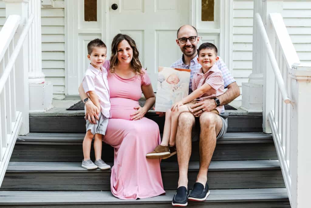 Nicole sits on a set of steps in a pink dress. Her husband and two sons are sitting with her. They are holding a picture of their baby who passed away.