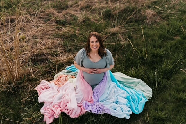 Clare is sitting on the ground holding her pregnant belly. The rainbow skirt is spread around her.