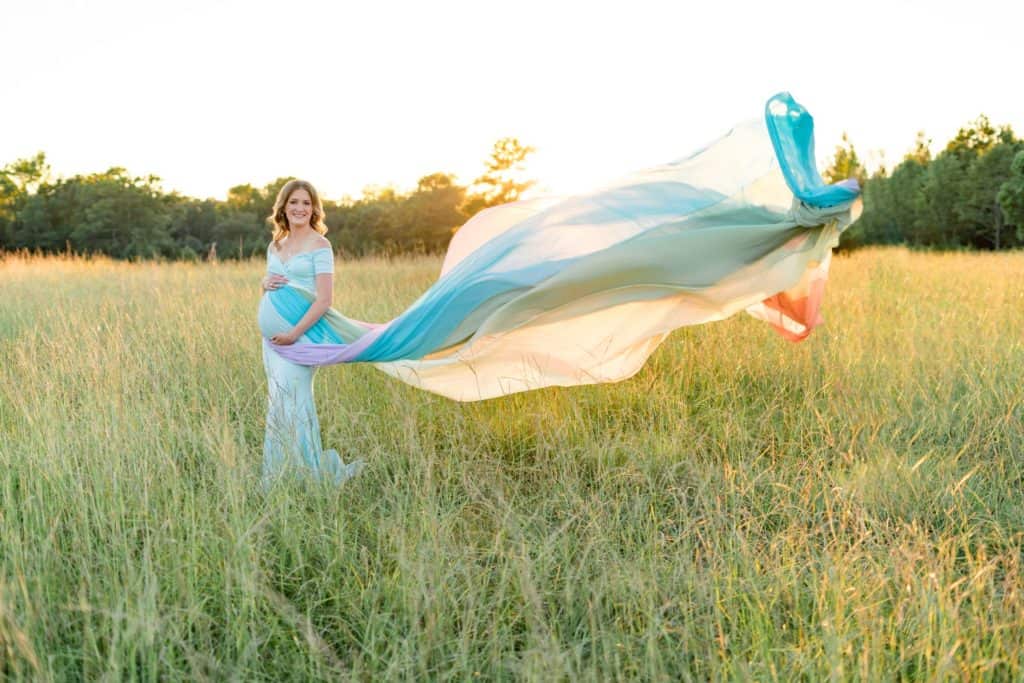 Kendall standing in a field of grass with the sun shining in the background. She is wearing a rainbow skirt that is flowing out behind her.