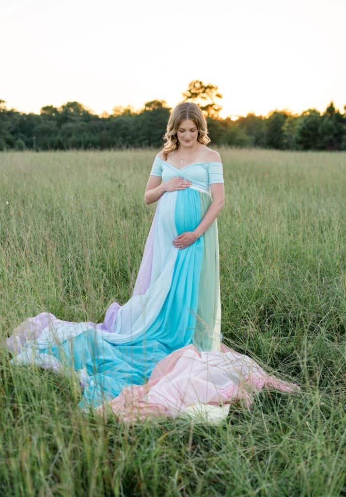 Kendall standing in a field of grass with trees and the sun shining in the background. She is wearing a rainbow skirt and holding her hands on her pregnant belly.