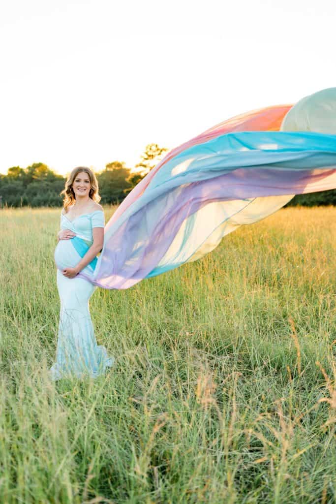Kendall standing in a field of grass with the sun shining in the background. She is wearing a rainbow skirt that is flowing out behind her.