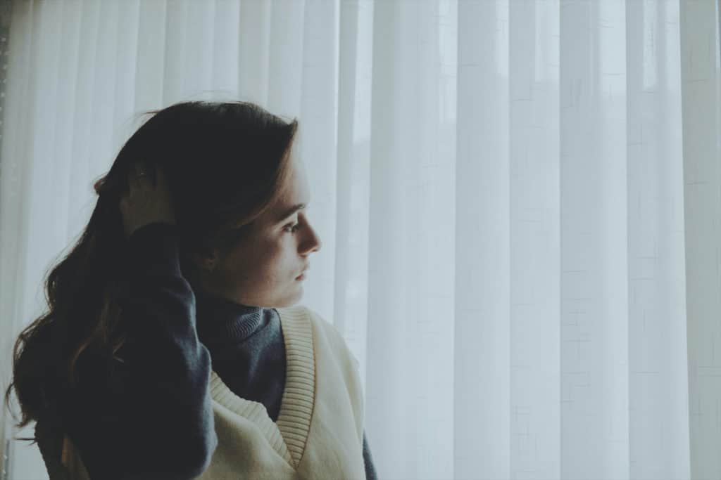 Woman is standing in front of white curtains looking towards the side. She looks worried.