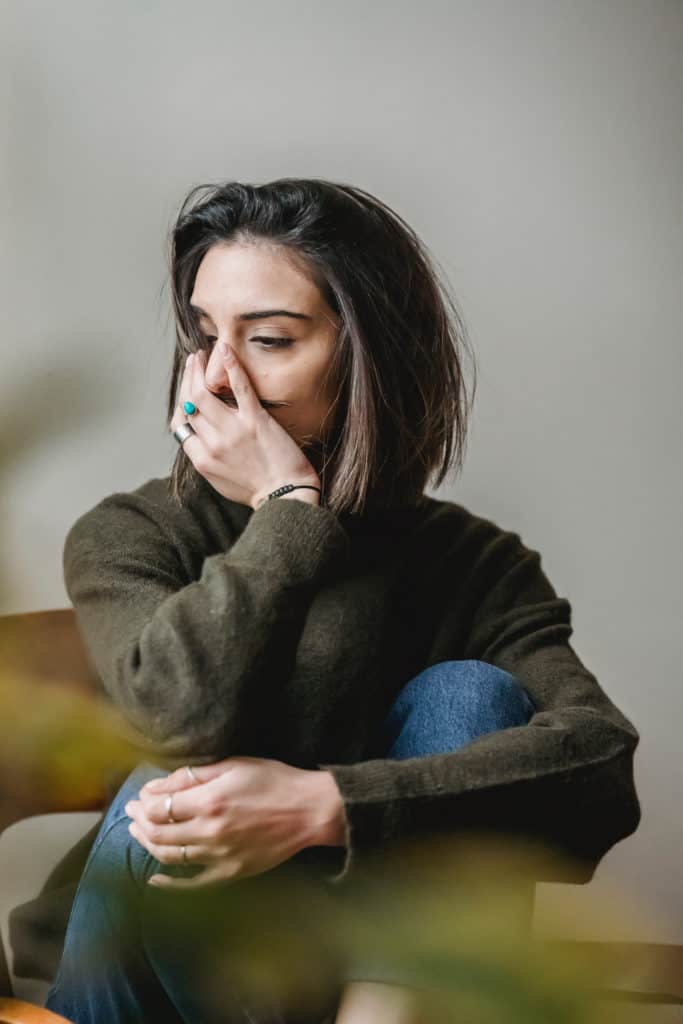 A woman sits with her hands around her knees looking worried.