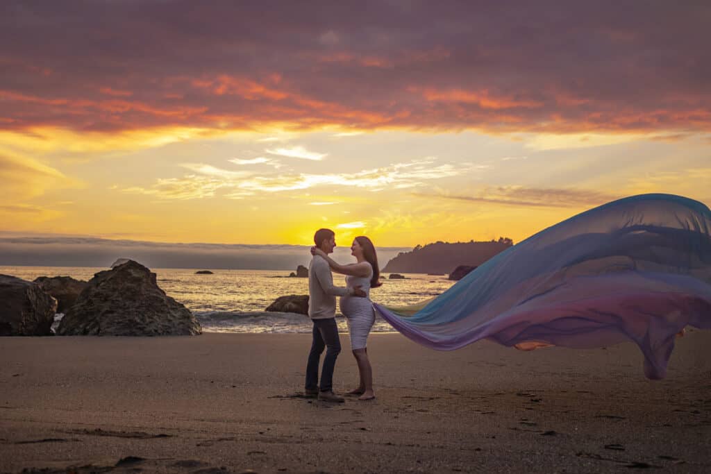 Alanna wears a white dress and the rainbow skirt.   The skirt flows out behind her.  Haru and Alanna stand on the beach with the sun setting.