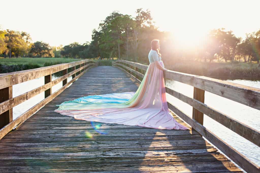 Rachel wears the rainbow skirt and stands on a bridge overlooking the water. The sun shines through the trees to the back of her.