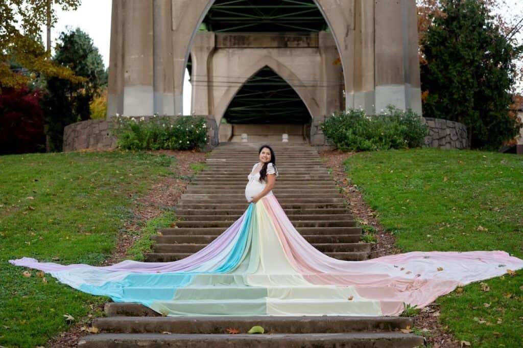 Briana wears a white dress and the rainbow skirt, which is spread out on the steps in front of her.