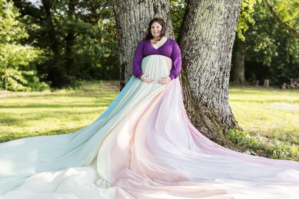 Kaycee wears a purple dress and the rainbow skirt. A large tree is behind her.