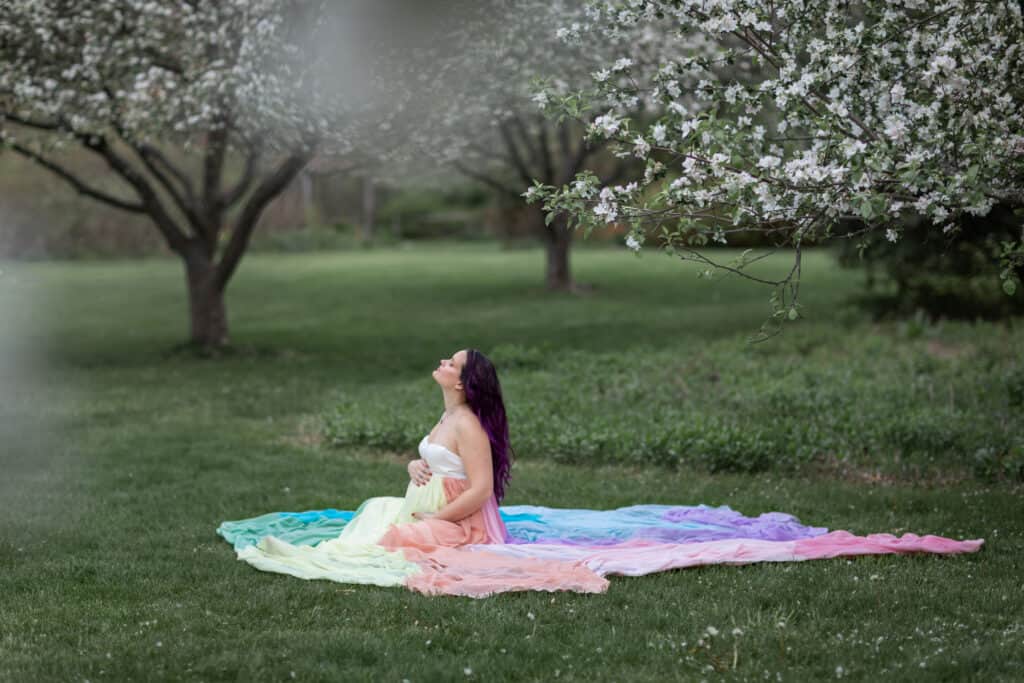 Kristina wears a white dress and the rainbow skirt. A tree with white flowers blooms behind her.