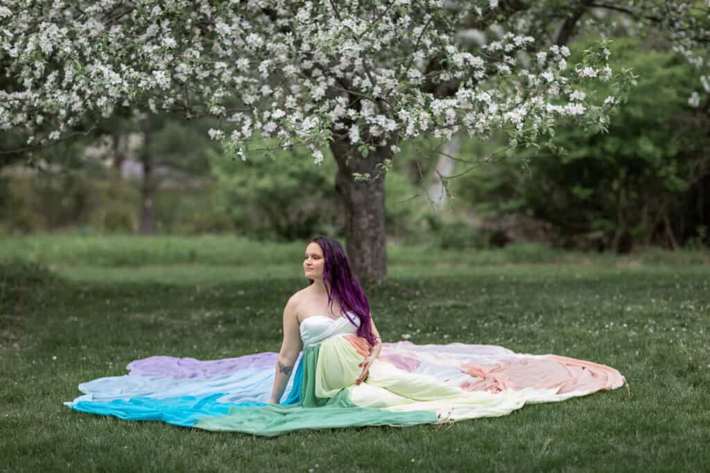 Kristina wears a white dress and the rainbow skirt. She sits on the ground with the skirt spread around her. A tree blooming with white flowers is behind her.
