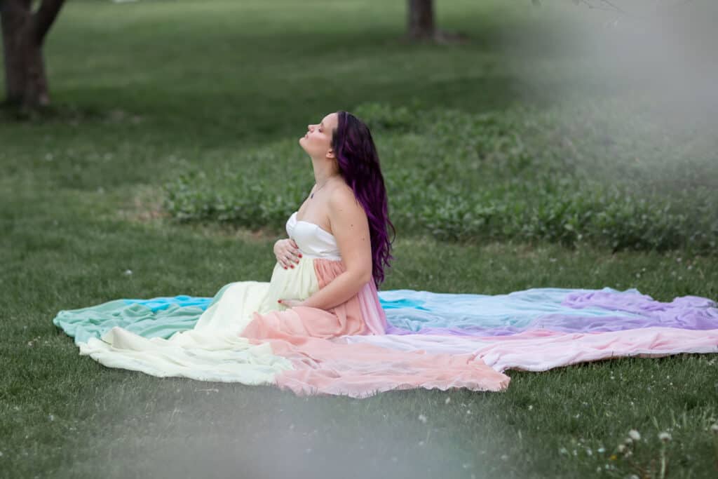 Kristina wears a white dress and the rainbow skirt. She sits on the ground and holds her pregnant belly.