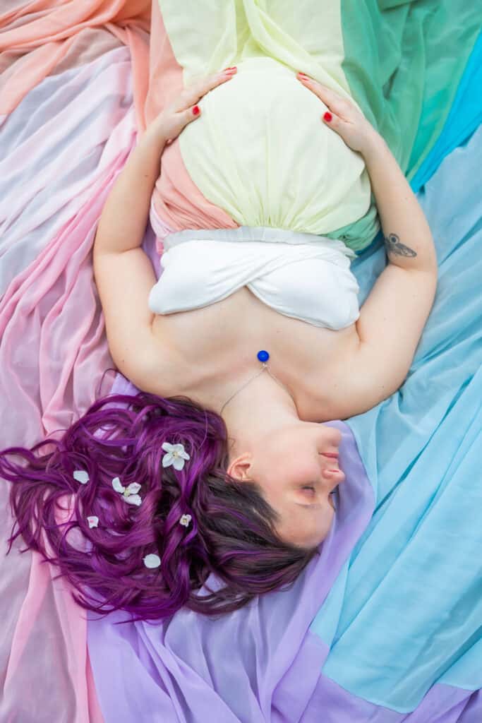 Kristina lays on top of the rainbow skirt. She has flowers in her hair and a blue charm on her necklace.