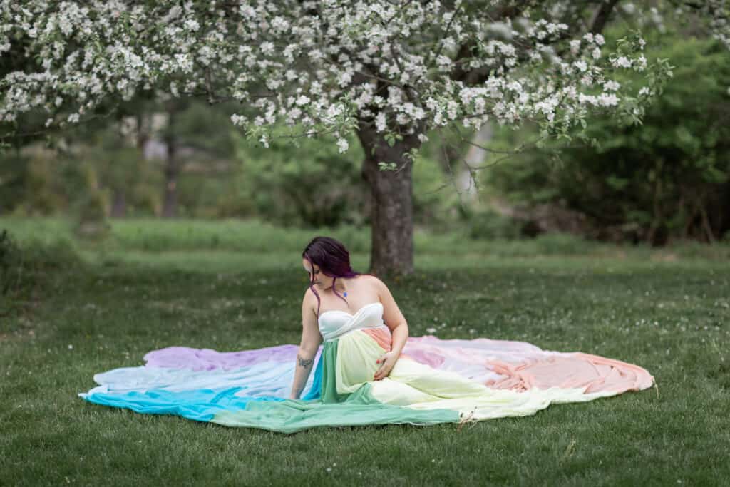 Kristina wears a white dress and the rainbow skirt. She sits on the ground with the skirt spread around her. She rests her hand on her pregnant belly.