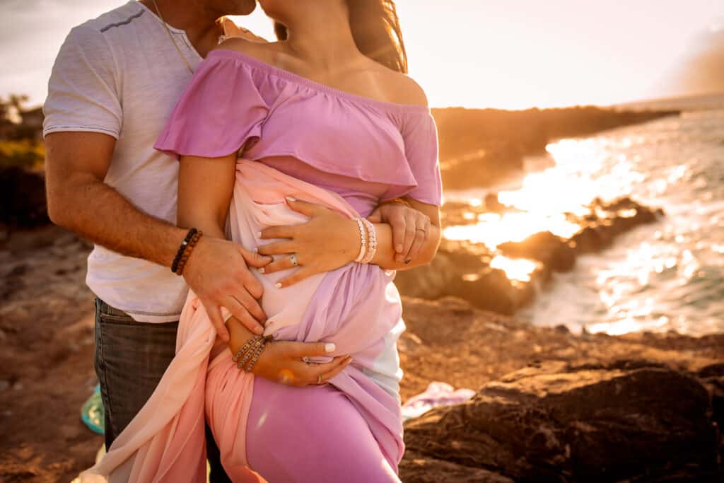 Rachael wears a pink dress and the rainbow skirt.  Her husband stands behind her and they rest their hands on her pregnant belly.