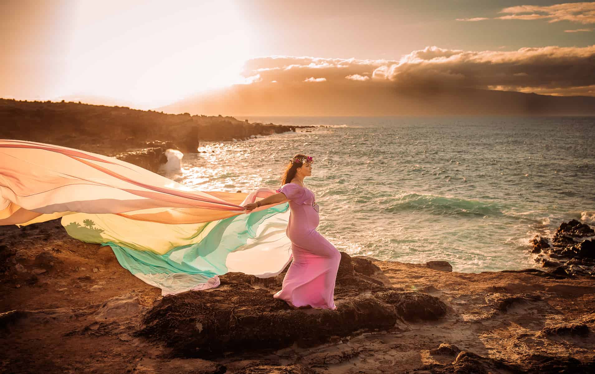 Rachael wears a pink dress and the rainbow skirt.   The skirt flows out behind her.  She stands in front of the ocean.