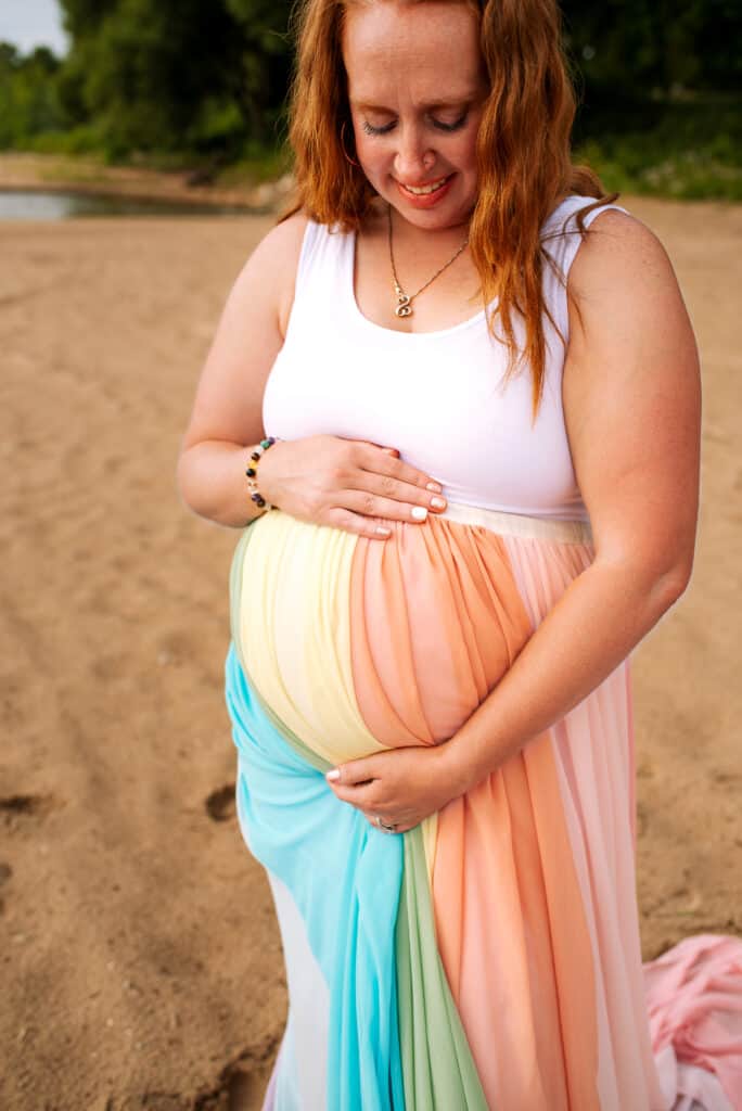 Ashlee wears a white dress and the rainbow skirt. She stands on the sand of a beach.