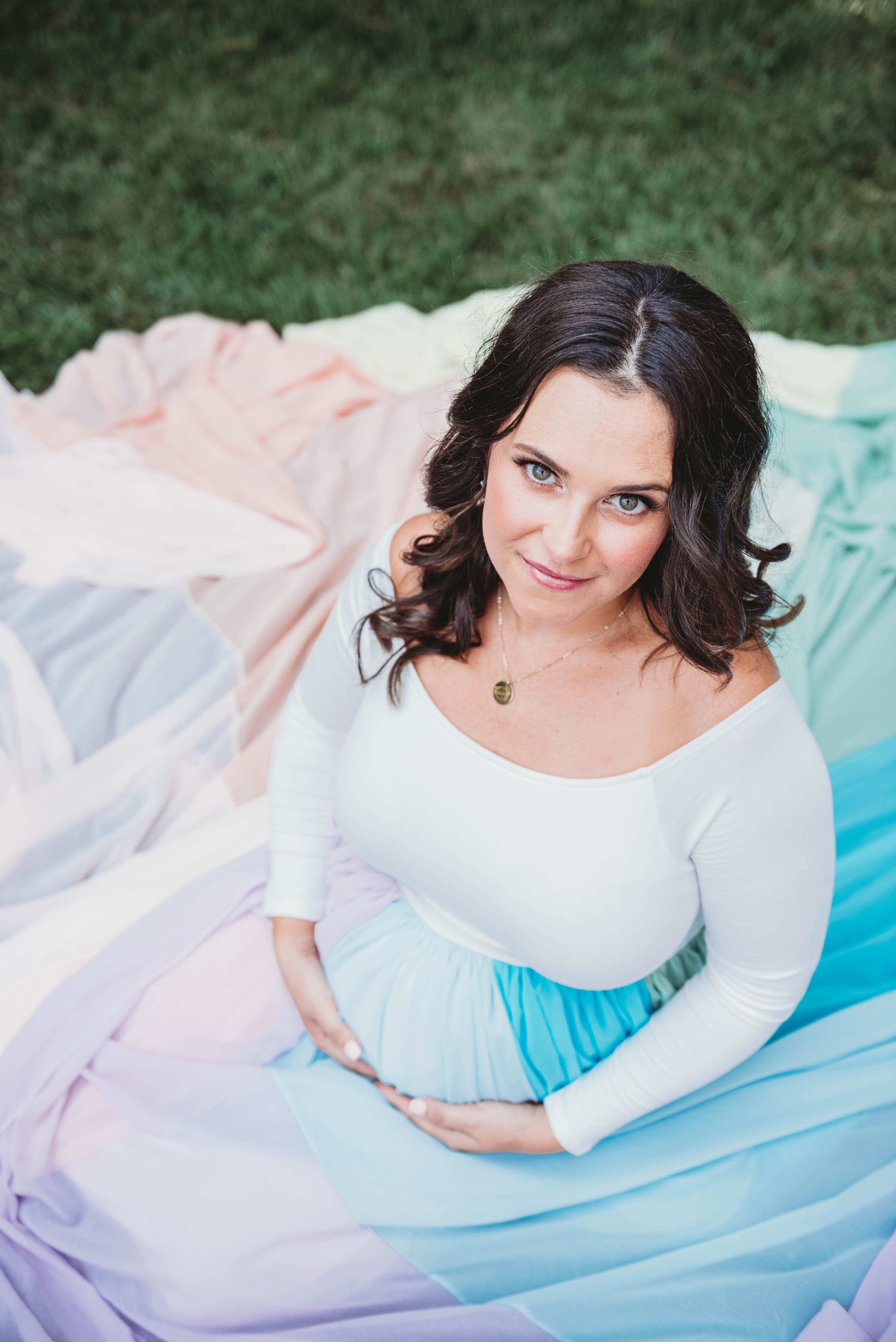 Molly wears a white dress and the rainbow skirt. She sits on the ground and holds her pregnant belly with both hands.