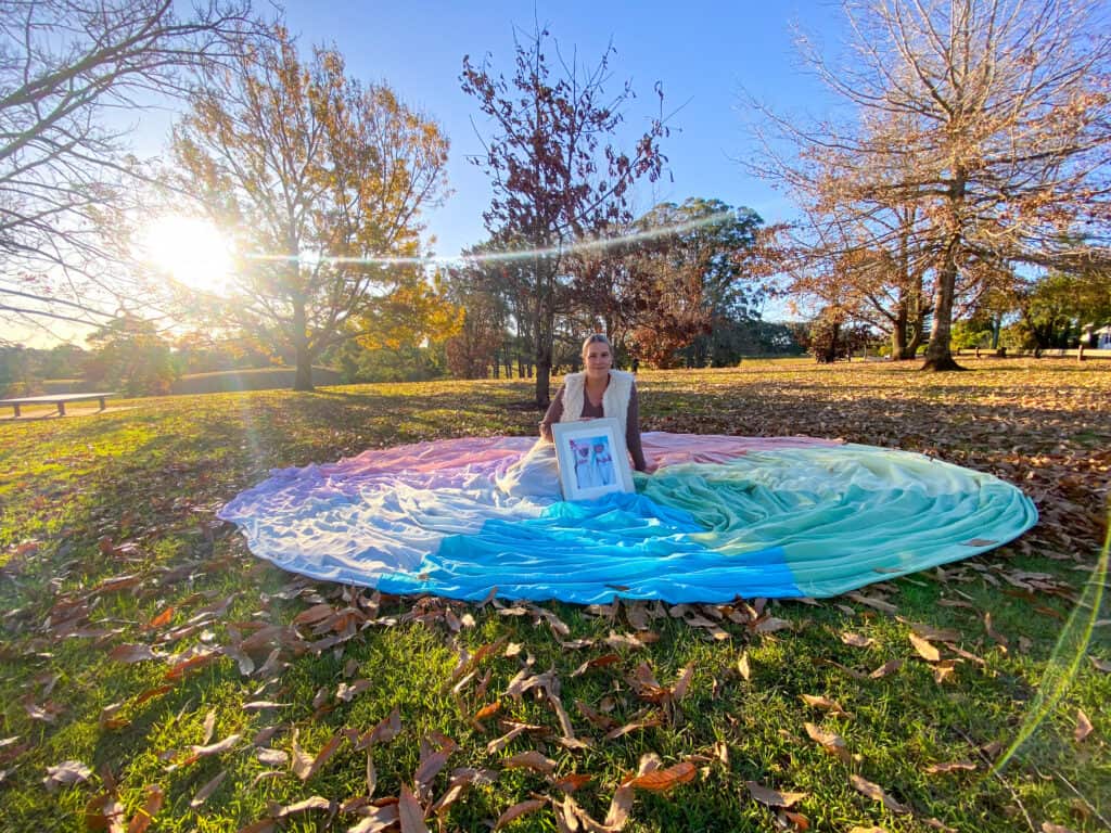 Samantha wears the rainbow skirt. She sits on the ground with the skirt spread around her on the ground. She holds a framed photo of her babies.