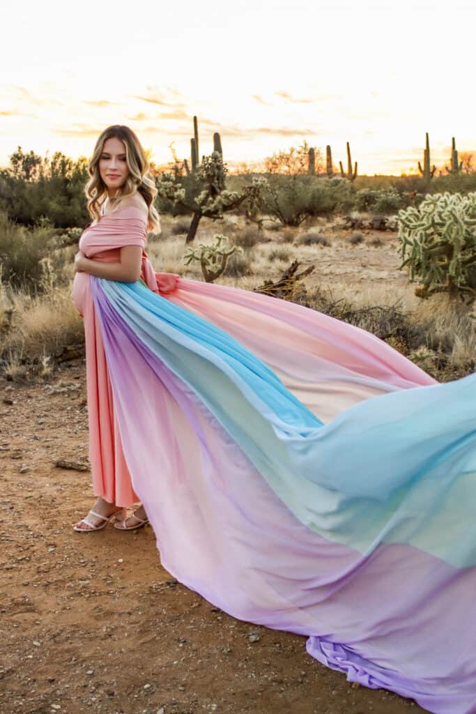 Ally wears a pink dress and the rainbow skirt. There are cacti in the background.