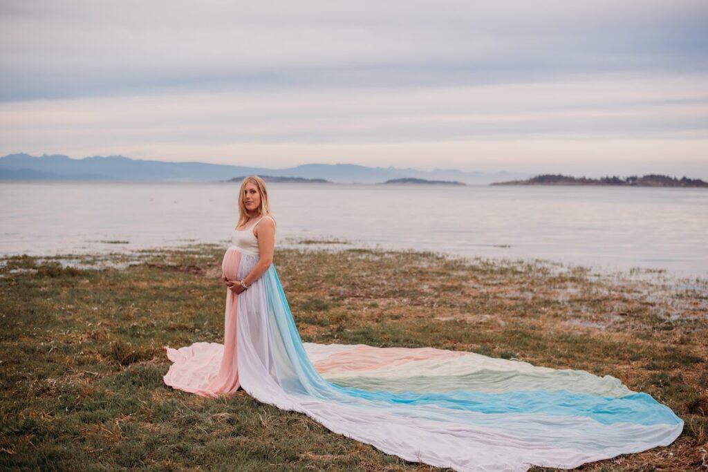 Erin wears a white dress and the rainbow skirt. She holds her pregnant belly with both hands.
