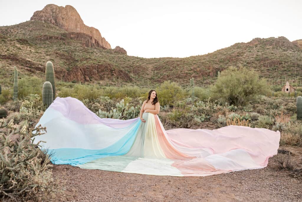 Alex wears the rainbow skirt. It is spread out all around here. There are hills and cacti in the background.