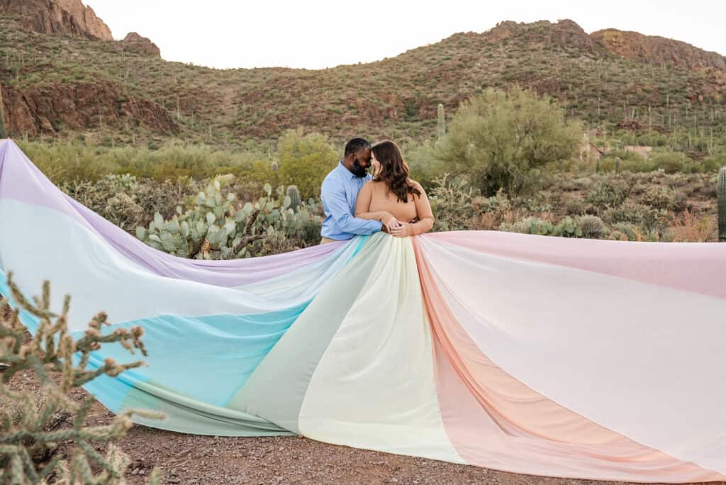 Alex wears the rainbow skirt. It is spread out all around here. There are hills and cacti in the background. Her husband stands next to her.