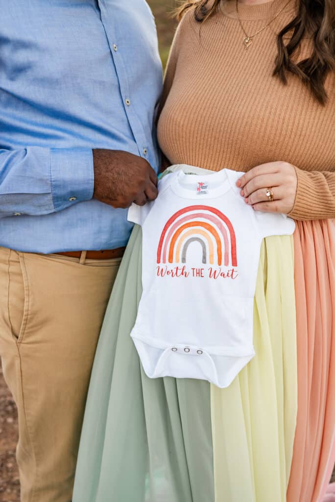 Alex wears the rainbow skirt. She and her husband each hold one side of a onesie that says "worth the wait" and has a rainbow.