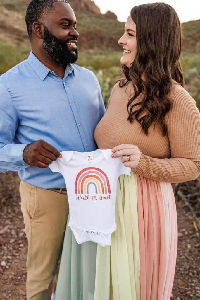 Alex wears the rainbow skirt. She and her husband each hold one side of a onesie that says "worth the wait" and has a rainbow.