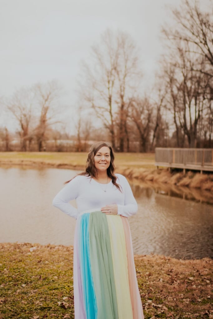 Kammie wears a white dress and the rainbow skirt. She stands in front of the water.