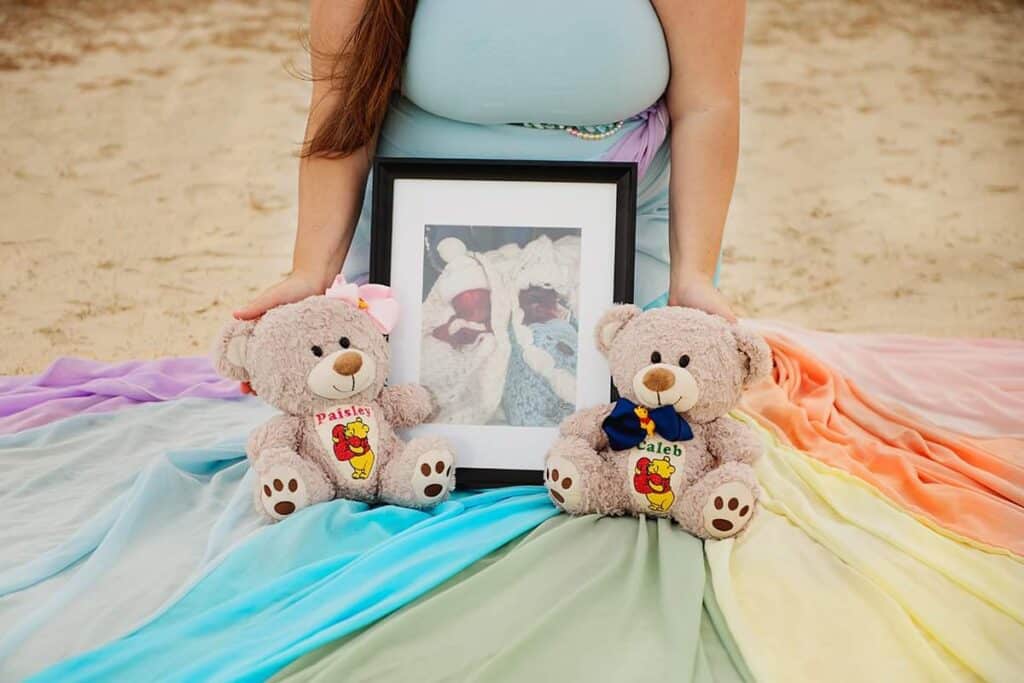Jessica wears a light blue dress with the rainbow skirt. On the skirt are two teddy bears with the names Paisley and Caleb. There is a framed photo of her two babies.