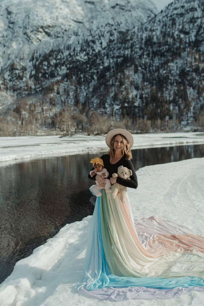 Jamie wears the rainbow skirt. She holds her daughter in one arm and a teddy bear in the other. There is snow on the ground and she stands next to the water.