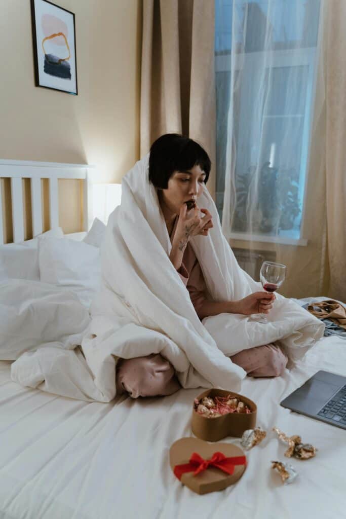A woman sits on the bed with a blanket wrapped around her. She is eating a piece of chocolate. There is a box of chocolates on the bed.
