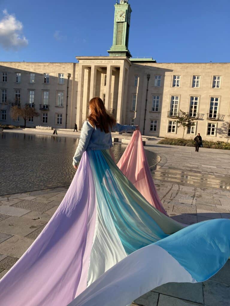 Poppy wears the rainbow skirt and holds it out to one side. A large town hall building is in the background.