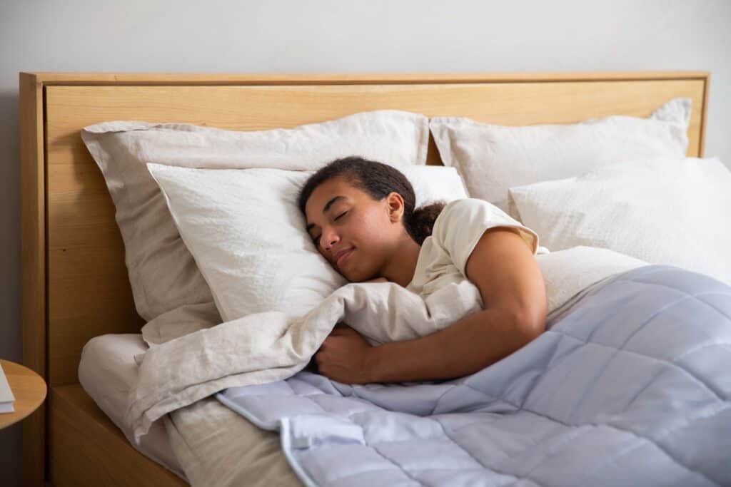 A woman sleeps in bed covered by a light blue weighted blanket.