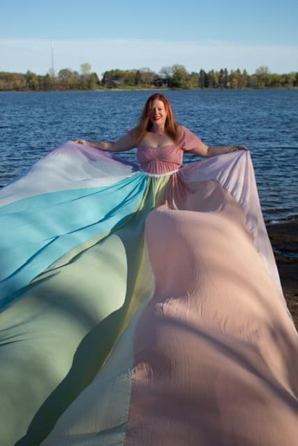 Alissa wears a pink dress and the rainbow skirt. She stands in front of the water holding the skirt up on both sides.