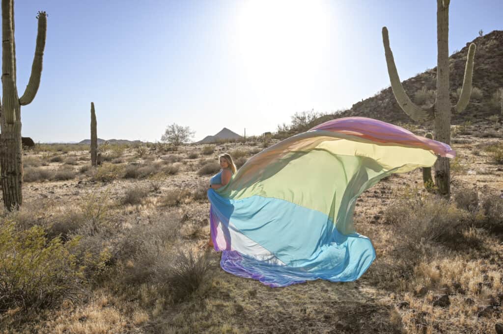Brittany wears a blue dress and the rainbow skirt. She stands in front of large cacti.