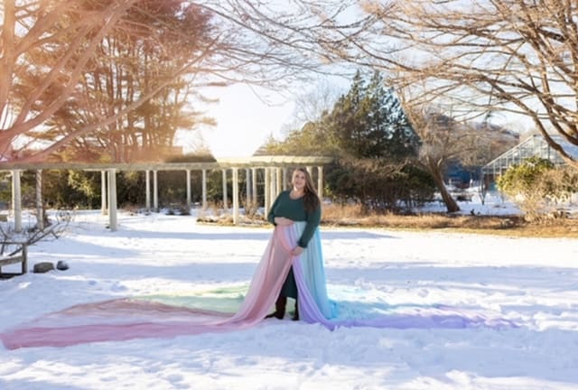 Heather wears a green dress and the rainbow skirt. She stands on the snow with trees in the background.