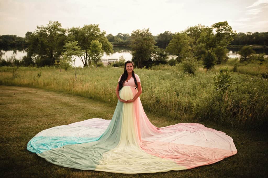 Arielle wears a pink dress and the rainbow skirt. She stands in front of tall green grass and trees.