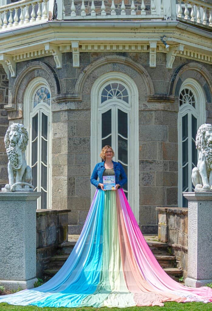 Julie wears the rainbow skirt and stands on a set of steps. She holds a copy of the book she wrote "Coral Couldn't Stay"