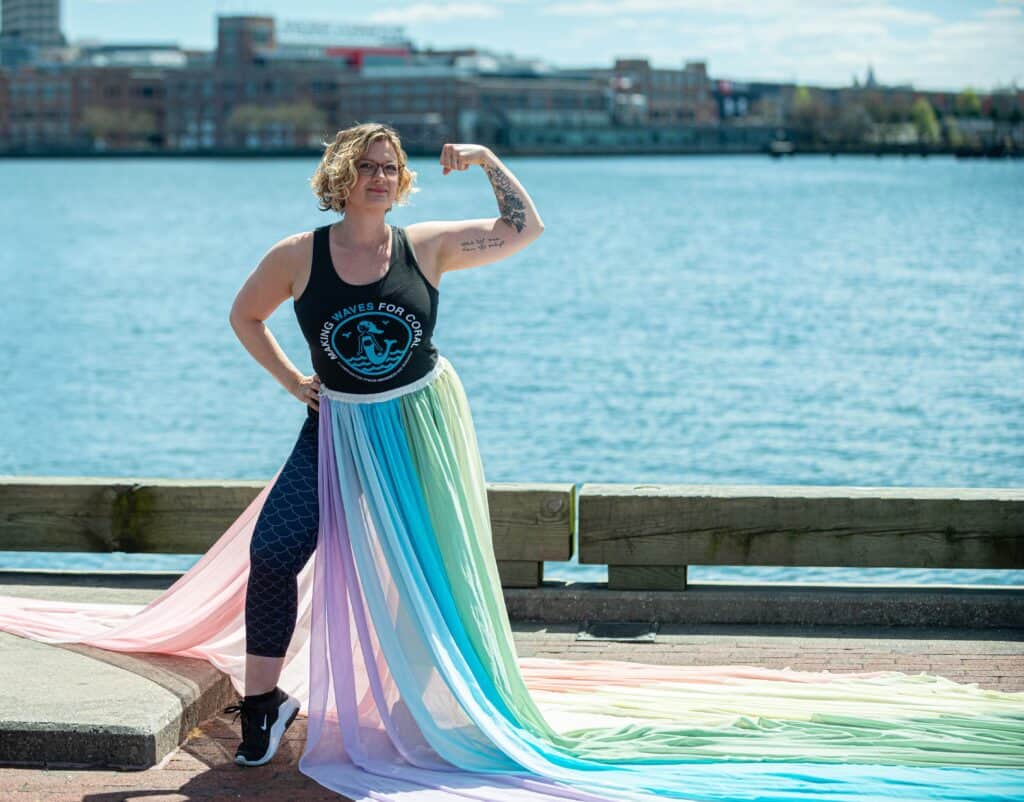 Julie wears the rainbow skirt and a shirt that says "Making Waves for Coral". She stands in front of the water.
