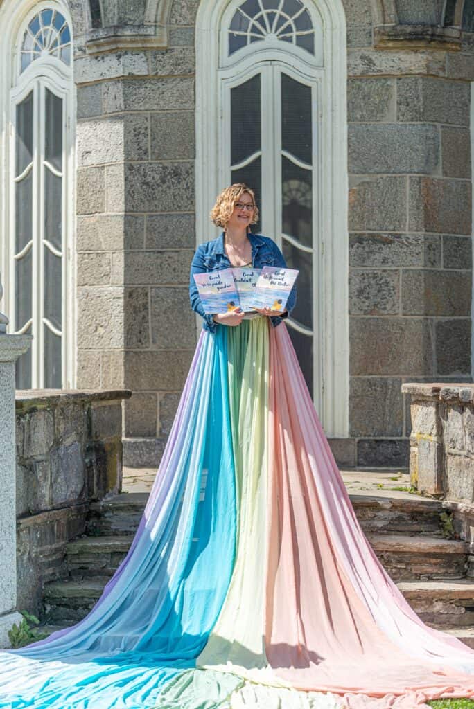 Julie wears the rainbow skirt. She holds three copies of the book she wrote called "Coral Couldn't Stay".