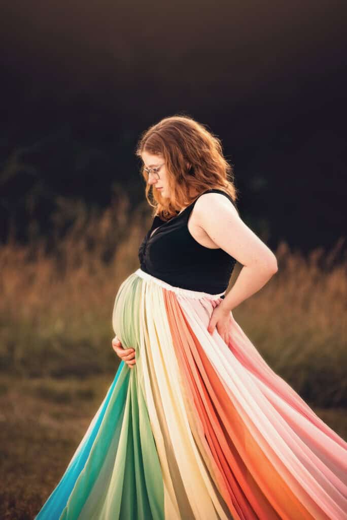 Ashton wears a black dress and the rainbow skirt. She holds her pregnant belly with one hand.