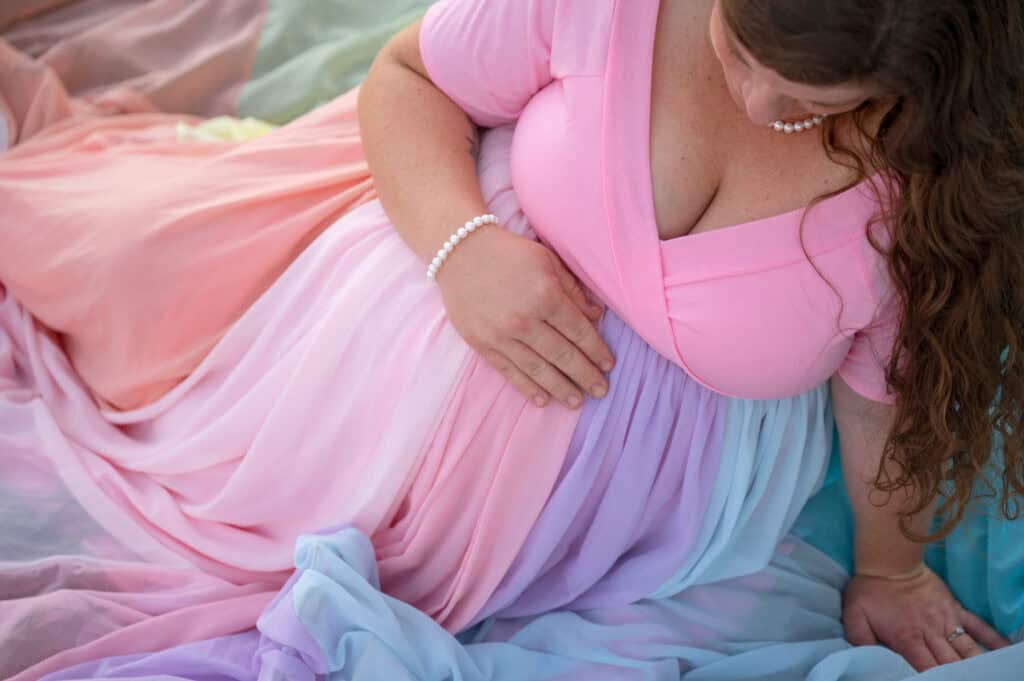 Rachael wears a pink dress and the rainbow skirt. She holds a hand on her pregnant belly.