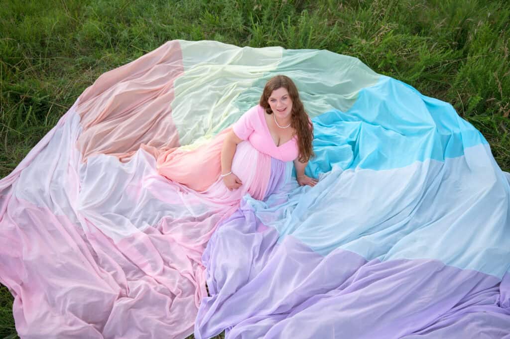 Rachael wears a pink dress and the rainbow skirt. She sits on the ground with the skirt spread around her on the ground.
