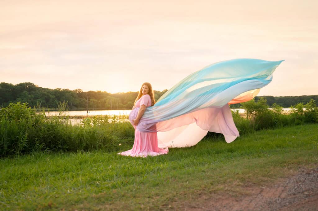Rachael wears a pink dress and the rainbow skirt. The skirt flows out in the air behind her. She stands in front of the water.